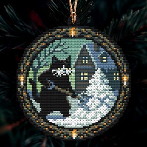 Op de afbeelding: Ronde kerstversiering met een zwarte kat in een besneeuwde sc&egrave;ne met een huis en een kerstboom. De versiering heeft een pixelachtig ontwerp, een donkere rand en een gouden ophangkoord.