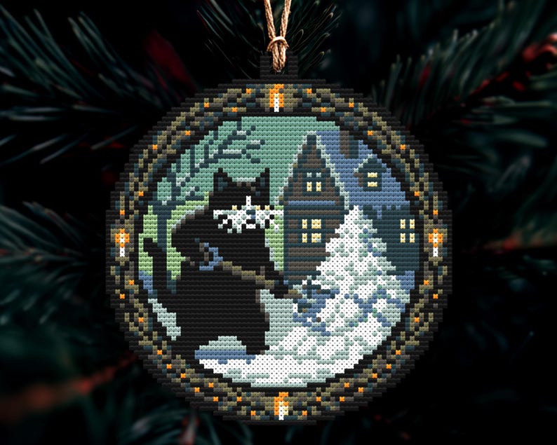 Op de afbeelding: Ronde kerstversiering met een zwarte kat in een besneeuwde sc&egrave;ne met een huis en een kerstboom. De versiering heeft een pixelachtig ontwerp, een donkere rand en een gouden ophangkoord.