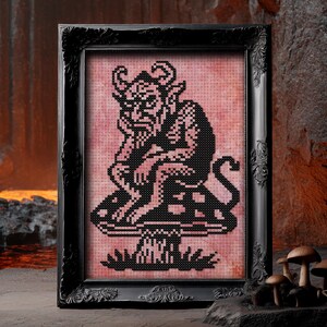May include: A framed cross-stitch artwork featuring a black devil figure with horns, sitting on a mushroom. The artwork is set against a pink background and framed in a black ornate frame. The artwork is displayed on a stone surface.