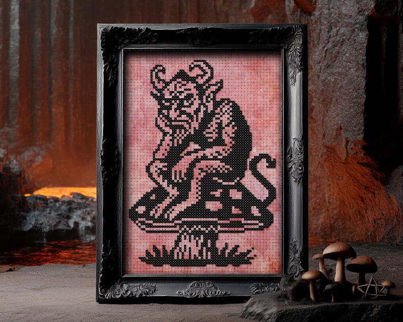 May include: A framed cross-stitch artwork featuring a black devil figure with horns, sitting on a mushroom. The artwork is set against a pink background and framed in a black ornate frame. The artwork is displayed on a stone surface.