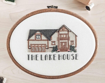 May include: An oval embroidery hoop featuring a cross-stitch design of a house. The house has a dark grey roof, beige siding, and brown trim. The text "THE LAKE HOUSE" is stitched below the house. The hoop is made of wood.