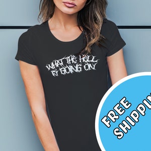 Può includere: Maglietta nera con testo bianco che recita "WHAT THE HELL IS GOING ON" in un font metallico. Un cerchio blu con testo bianco che recita "SPEDIZIONE GRATUITA!" si trova nell'angolo in basso a destra dell'immagine.
