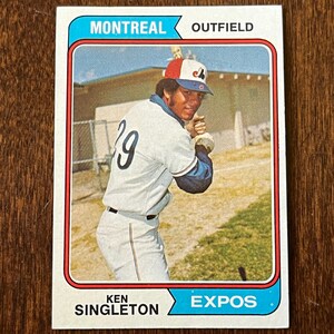 Puede incluir: Una tarjeta de béisbol vintage de Ken Singleton de los Montreal Expos. La tarjeta muestra al jugador con uniforme blanco con el número 29, una gorra roja, blanca y azul, y un bate. El texto incluye MONTREAL, OUTFIELD y EXPOS.
