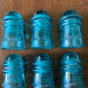 Op de afbeelding: Zes blauwe glazen isolatorknoppen met de tekst "Hemingray" op elk. De isolatoren zijn in verschillende maten en hebben een getextureerd oppervlak.