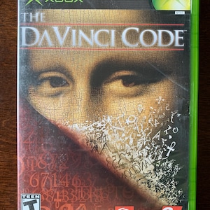 May include: An Xbox video game case for The DaVinci Code featuring a close-up of the Mona Lisa's eyes with a cracked texture effect. The cover includes the title, ESRB Teen rating, Columbia Pictures, The Collective, and 2K logos on a red background.