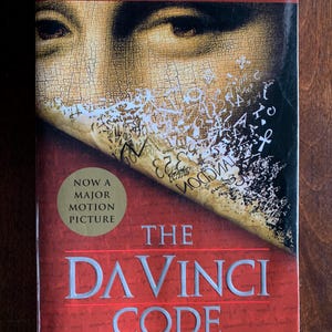 Op de afbeelding: Een paperback getiteld The Da Vinci Code van Dan Brown met een rode kaft met een close-up van een gezicht en verspreide symbolen. Een gouden ronde sticker op de kaft vermeldt Now a Major Motion Picture en de tekst The #1 Worldwide Bestseller.