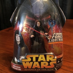 May include: A Star Wars action figure of Emperor Palpatine from the Revenge of the Sith film. The figure is in a plastic blister pack with a red and orange background. The figure is wearing a black robe and has a red lightsaber. The packaging includes the text "Firing Force Lightning".