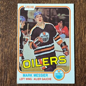 May include: Vintage O-Pee-Chee hockey card featuring Mark Messier of the Oilers. The card has a white border and displays the Oilers logo. Messier is wearing a black and orange jersey with the number 11. The text "OILERS" is prominently displayed.