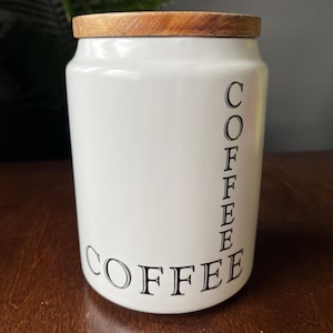 Puede incluir: Un bote de café de cerámica blanca con tapa de madera. La palabra "COFFEE" está impresa vertical y horizontalmente en negro, centrada en el bote. El bote es cilíndrico y se encuentra sobre una superficie de madera oscura.