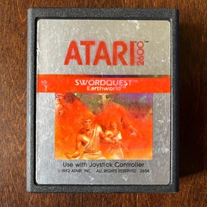 May include: A silver Atari 2600 game cartridge with a red label. The label features the text "ATARI 2600", "SWORDQUEST™", "Earthworld™", and "Use with Joystick Controller". The label also includes the copyright information "© 1982 ATARI, INC. ALL RIGHTS RESERVED 2656".