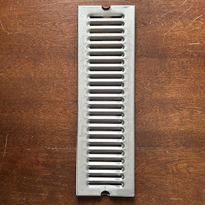 May include: A rectangular, silver-colored vent cover with a series of horizontal slots. The vent cover has a slightly rounded top and bottom edge, and is set against a dark brown wooden surface. The vent cover is designed for air circulation.