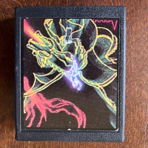 Può includere: Una cartuccia di videogioco vintage con una custodia in plastica nera e un'immagine colorata di un drago verde con accenti rossi e viola luminosi.