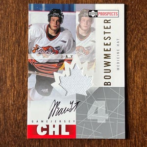 Op de afbeelding: Een hockey trading card met een speler in een wit en oranje shirt. De kaart heeft een esdoornblad ontwerp en de naam "Bouwmeester". De tekst "Game Jersey CHL" is ook zichtbaar. De kaart is rechthoekig van vorm.