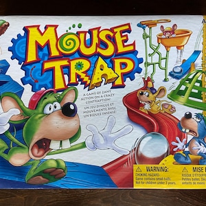 Peut inclure: Une boîte de jeu de société colorée pour Mouse Trap, avec un dessin animé d'une souris qui s'enfuit d'un piège complexe. La boîte comporte un avertissement concernant les risques d'étouffement en raison de petites billes.