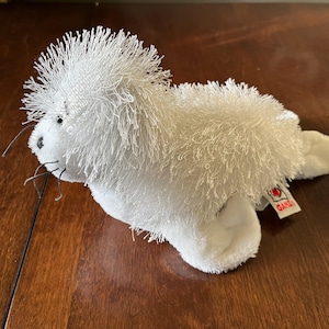 Puede incluir: Un juguete de peluche de foca blanca con una melena y bigotes desgreñados. El peluche tiene una etiqueta que dice "GANZ". La foca está tumbada sobre una superficie de madera.