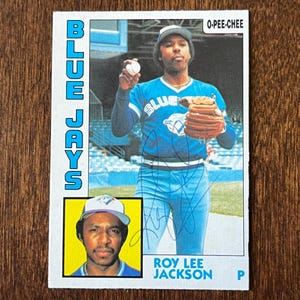 Puede incluir: Una tarjeta de béisbol vintage O-Pee-Chee con Roy Lee Jackson de los Blue Jays. La tarjeta muestra a Jackson con su uniforme, sosteniendo una pelota y un guante de béisbol. La tarjeta tiene una pequeña foto insertada y las palabras "Blue Jays" en azul.