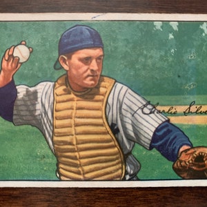 May include: A vintage baseball card featuring a colour illustration of a baseball player wearing a blue cap, a striped jersey, and a catcher's chest protector. The player is throwing a white baseball with his right hand. The card has a green background and the player's name, "Charlie Silvera", is handwritten in black ink in the bottom right corner.