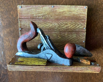 May include: A wooden shelf with a silver and brown hand plane, a ruler, and a small wooden tool. The shelf is mounted on a wooden background.
