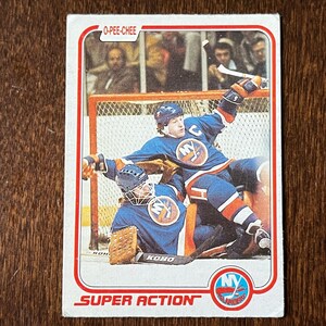 May include: O-Pee-Chee hockey card featuring New York Islanders players in action. The card has a red border and the words "SUPER ACTION" at the bottom. The players are wearing blue and orange jerseys. The goalie is wearing a brown glove.