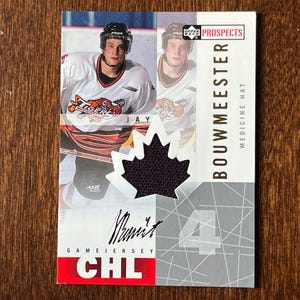 Op de afbeelding: Een hockey trading card met een speler in een wit en oranje shirt met de tekst "Tigers". De kaart bevat een zwart esdoornblad patch en de naam "Bouwmeester". De tekst "Game Jersey CHL" is zichtbaar.