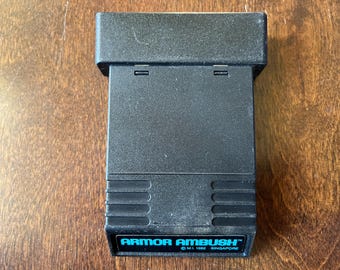 May include: Black cartridge for the Nintendo Entertainment System (NES) video game console. The cartridge has a label that reads "Armor Ambush" with the copyright information "C M.L 1982 SINGAPORE".