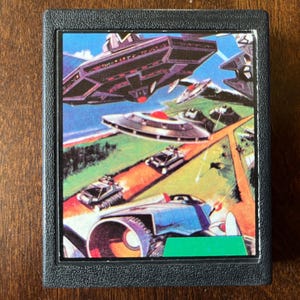 Può includere: Una cartuccia di videogioco vintage con un'illustrazione colorata di astronavi e veicoli su un paesaggio verde e marrone. La cartuccia è nera con un'etichetta verde.