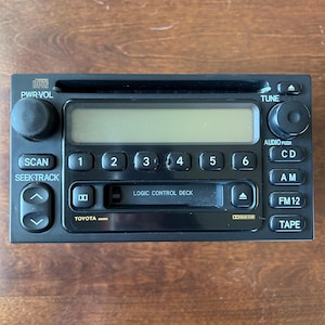 Peut inclure: Un autoradio Toyota noir comprenant une fente pour lecteur CD, un lecteur de cassette avec contrôle logique, un écran numérique et des boutons marqués PWR-VOL, TUNE, SCAN, SEEK-TRACK, CD, AM, FM1-2 et TAPE, avec logos Dolby B NR et Compact Disc.