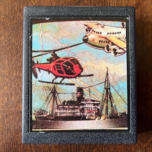 Puede incluir: Una caja de juego vintage que presenta un helicóptero rojo volando sobre un gran barco con un avión blanco en el fondo. La escena se desarrolla en un cielo tormentoso con rayos.