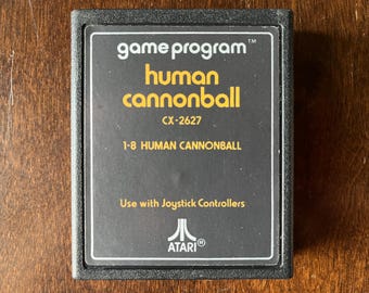 May include: Black Atari game cartridge with yellow text that reads "game program human cannonball CX-2627 1-8 HUMAN CANNONBALL TM Use with Joystick Controllers ATARIⓇ".