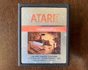 May include: A vintage Atari 2600 game cartridge for Vanguard. The cartridge is silver with a red label that says "Vanguard" and features a spaceship in a futuristic setting.