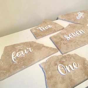 May include: Five beige marble tiles with white cursive numbers on them. The numbers are "one", "two", "four", "seven", and "eight".