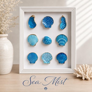 May include: A white rectangular frame containing nine individual seashells arranged in a three-by-three grid. Each shell is painted in various shades of blue with gold-painted edges. The text "Sea Mist" is printed in blue script below the frame.