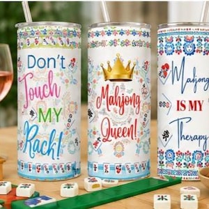 Könnte beinhalten: Drei weiße isolierte Becher mit Strohhalmen, jeweils mit einem Mahjong-Thema und Text. Einer sagt "Don't Touch My Rack!", ein anderer "Mahjong Queen!" und der dritte "Mahjong is My Therapy". Mahjong-Steine und ein Glas Wein sind im Hintergrund.