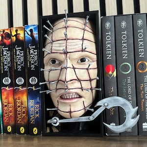 May include: A display of books, including the Percy Jackson series and The Lord of the Rings trilogy, on a wooden shelf. A sculpture of a face with nails and a metal hook is placed among the books.