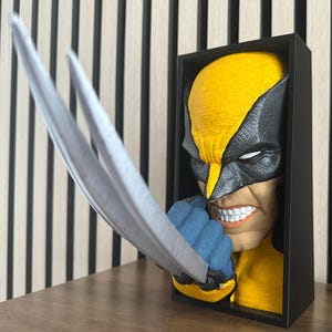 May include: A Wolverine bust sculpture featuring the character's face with yellow and black mask, blue gloves, and silver claws. The sculpture is set in a black frame, with the claws extending out. The background is a wooden wall.