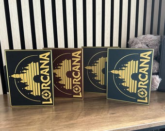 May include: Four black, brown, and gray square plaques with gold lettering and a gold graphic of a castle with a sun setting behind it. The text on the plaques reads "Lorcana".