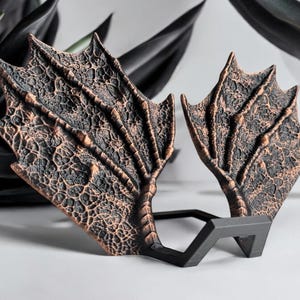 Op de afbeelding: Een paar decoratieve drakenvleugels gemaakt van koperkleurig metaal. De vleugels zijn gedetailleerd met een textuur, schubbenontwerp en zijn gemonteerd op een zwarte standaard.
