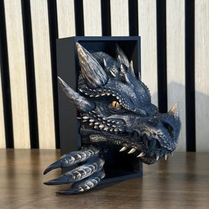 May include: A black dragon head sculpture with silver accents, peeking out of a black frame. The dragon has a fierce expression with sharp teeth and claws.