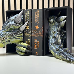 May include: A dragon bookend with a green and blue painted finish. The dragon's head is on the left side of the bookend, and its tail is on the right. The bookend is holding up two books with the titles "Iron Flame" and "Fourth Witch" by Rebecca Yarros.