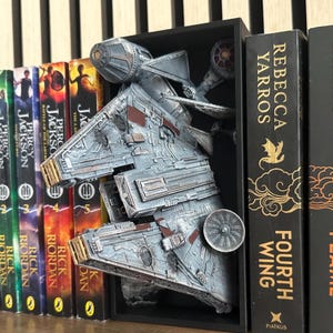May include: A detailed, grey Millennium Falcon model bookend, positioned between books. The bookend is in a black frame, with visible details like the cockpit and panels. Books include "Percy Jackson" and "Fourth Wing."