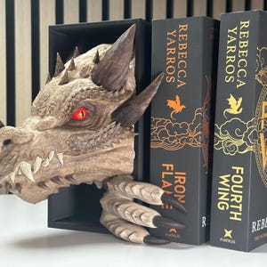 May include: A dragon bookend with a textured, brown and gray finish. The dragon has a red eye and sharp teeth. The bookend is holding up two books with black covers. The book covers have gold lettering that reads "Iron Flame" and "Fourth Wing" by Rebecca Yarros.