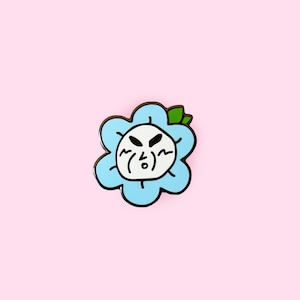 Op de afbeelding: Een blauwe emaille pin in de vorm van een bloem met een wit gezicht en een groen blad. De bloem heeft een cartoonachtige boze uitdrukking met zwarte ogen en een frons.