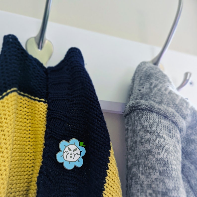 Op de afbeelding: Twee gebreide truien hangen aan haken. E&eacute;n trui is geel en blauw gestreept. De andere trui is grijs. Een blauwe emaille pin met een cartoon bloem gezicht is op de gestreepte trui.