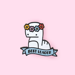May include: White enamel pin with a cartoon dinosaur wearing a flower crown and a blue banner that says "BEST LEADER".