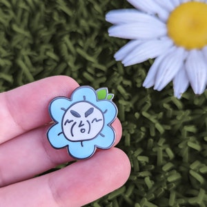 Op de afbeelding: Een blauwe emaille pin in de vorm van een bloem met een wit gezicht en zwarte ogen. De bloem heeft een groen blaadje bovenop. Het gezicht heeft een verraste uitdrukking met een kleine mond en grote ogen.