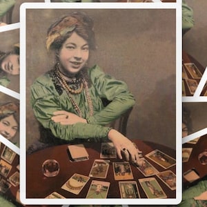May include: A woman wearing a green dress and a beaded necklace sits at a table with a crystal ball and a spread of tarot cards. The cards are face up and show various images and symbols.