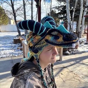Puede incluir: Un gorro de dragón de ganchillo hecho a mano en tonos azules, amarillos y verdes. El gorro presenta cuernos, un hocico y orejeras trenzadas. El gorro está diseñado para parecerse a la cabeza de un dragón.