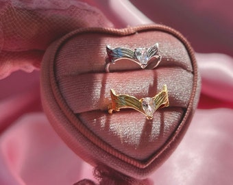 May include: Two rings with clear stones set in a heart-shaped ring box. The ring on top is silver with a clear stone and the ring on the bottom is gold with a clear stone.