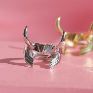 May include: Two rings with a horned helmet design, one silver and one gold, are displayed on a pink surface. The silver ring is in focus, showcasing the intricate details of the helmet's design. The gold ring is slightly blurred in the background.