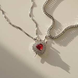 May include: Two silver chains with a pendant featuring a red heart with a crown and a silver sword. The pendant is adorned with small clear crystals.